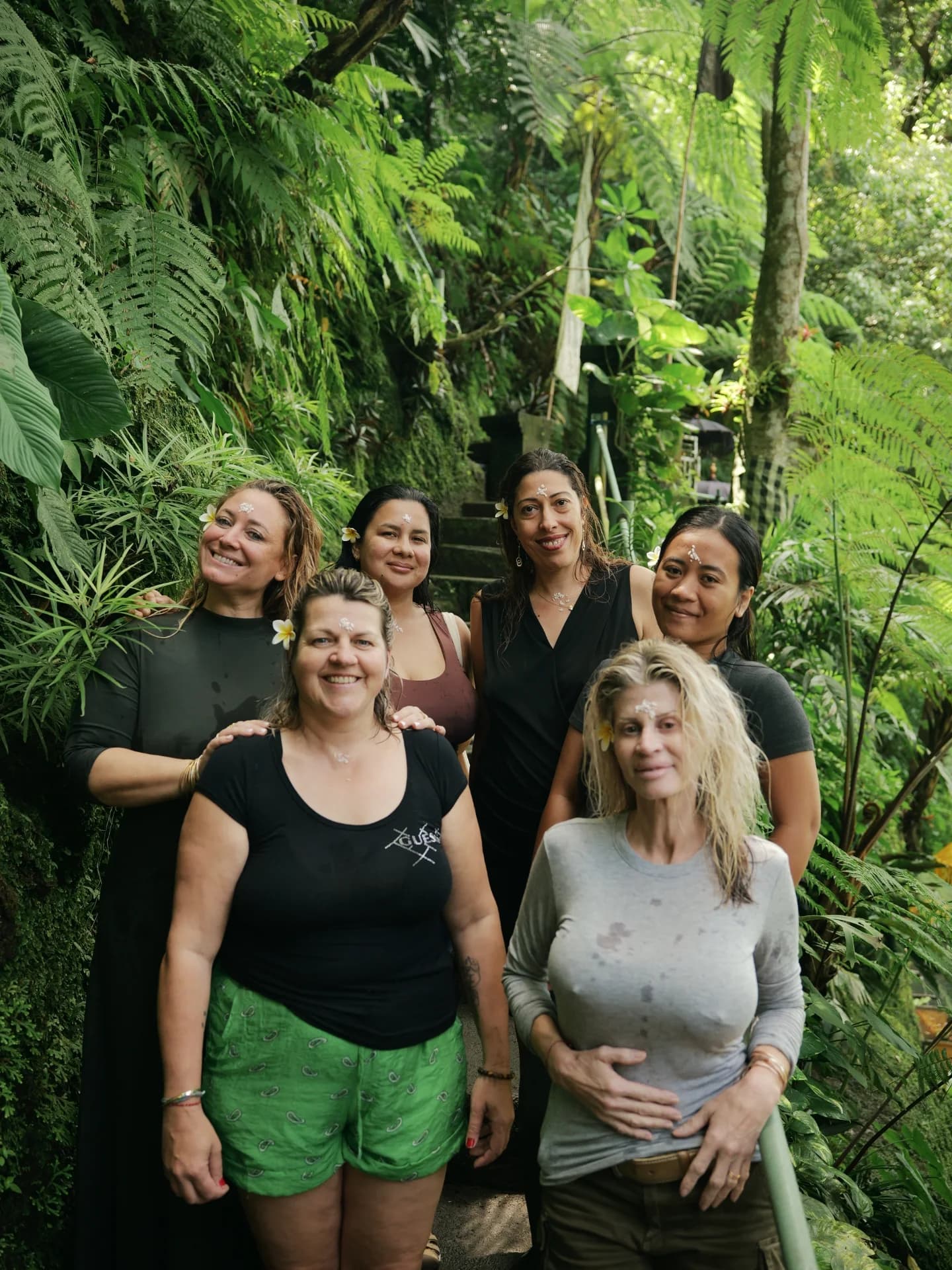 Six femmes souriantes posent dans une jungle luxuriante avec des fleurs et marques rituelles.
