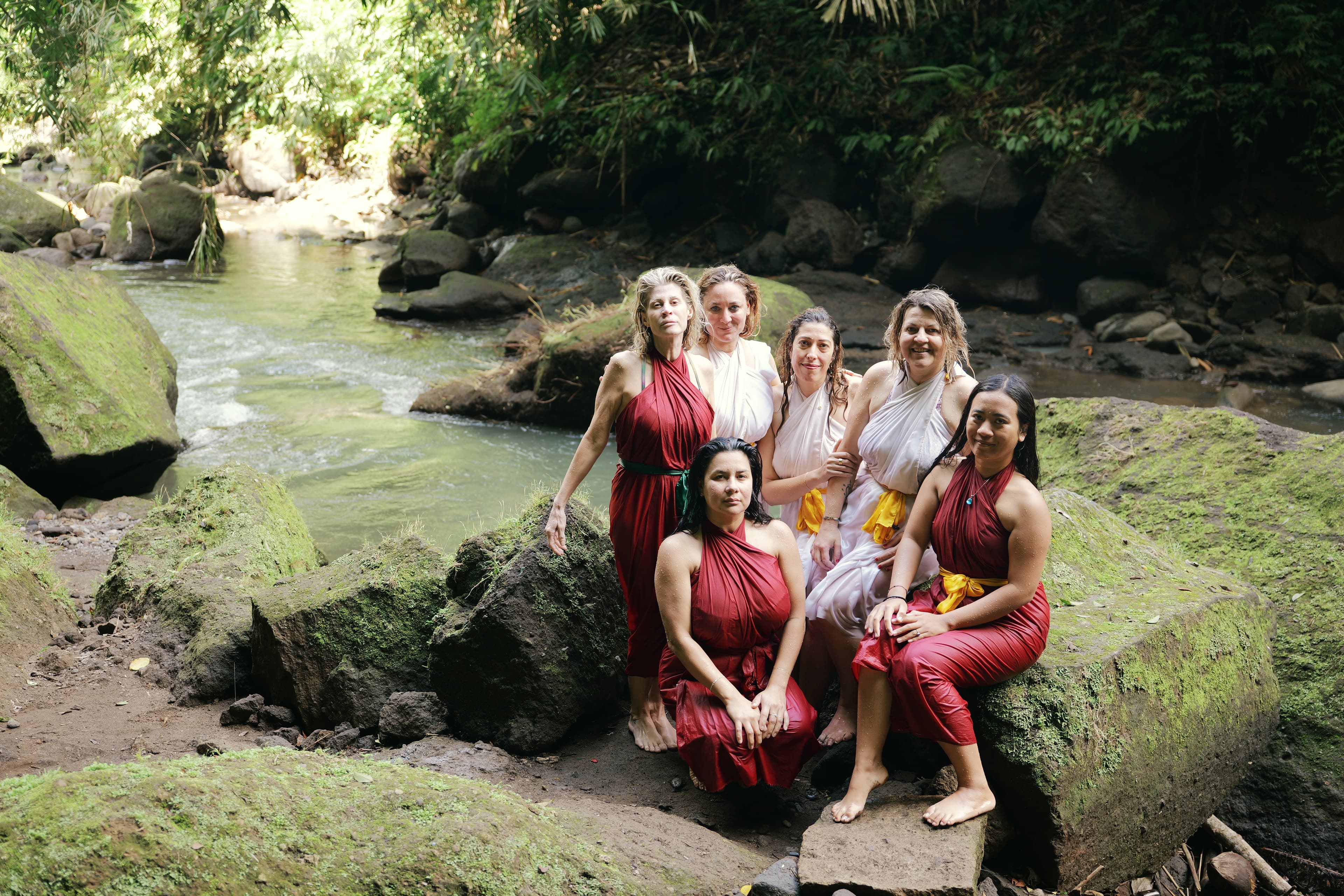 Six femmes en sarongs rouges et blancs posent sur des rochers près d'une rivière.