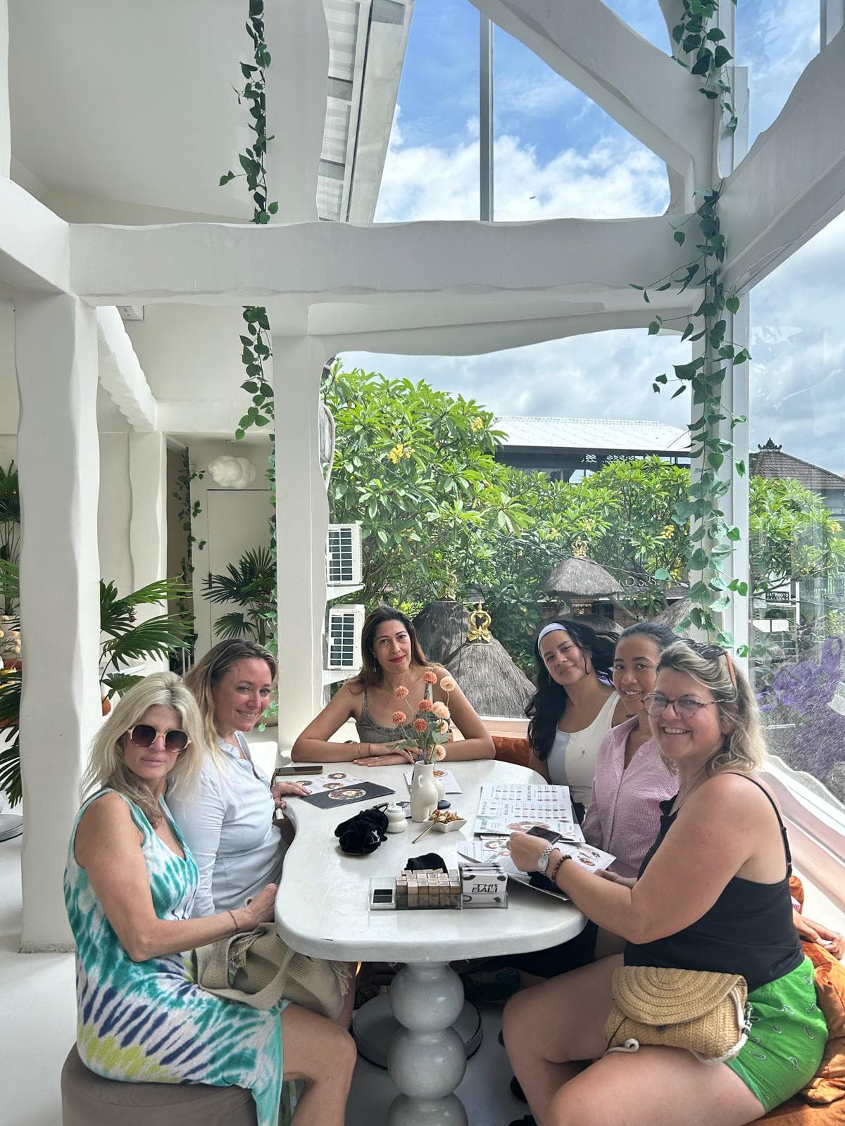 Six femmes souriantes attablées dans un restaurant blanc et lumineux entouré de plantes vertes.