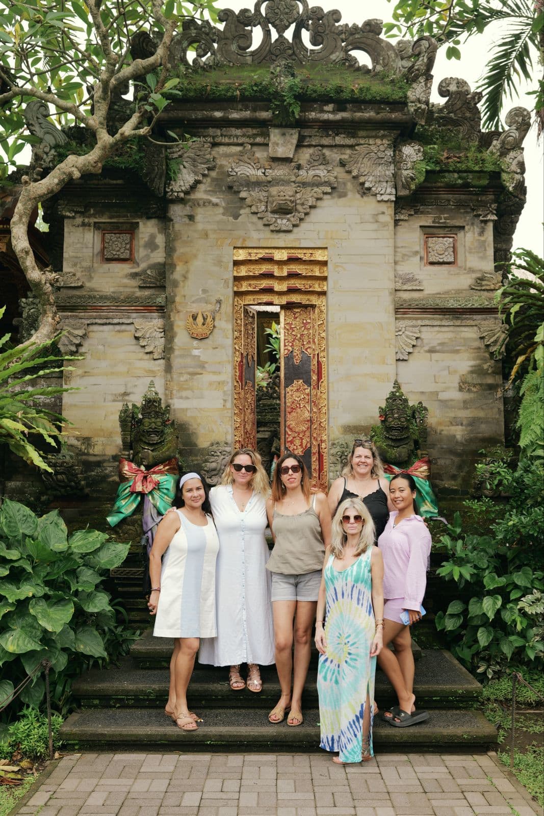 Six femmes posent devant une porte de temple balinais sculptée entourée de végétation luxuriante.