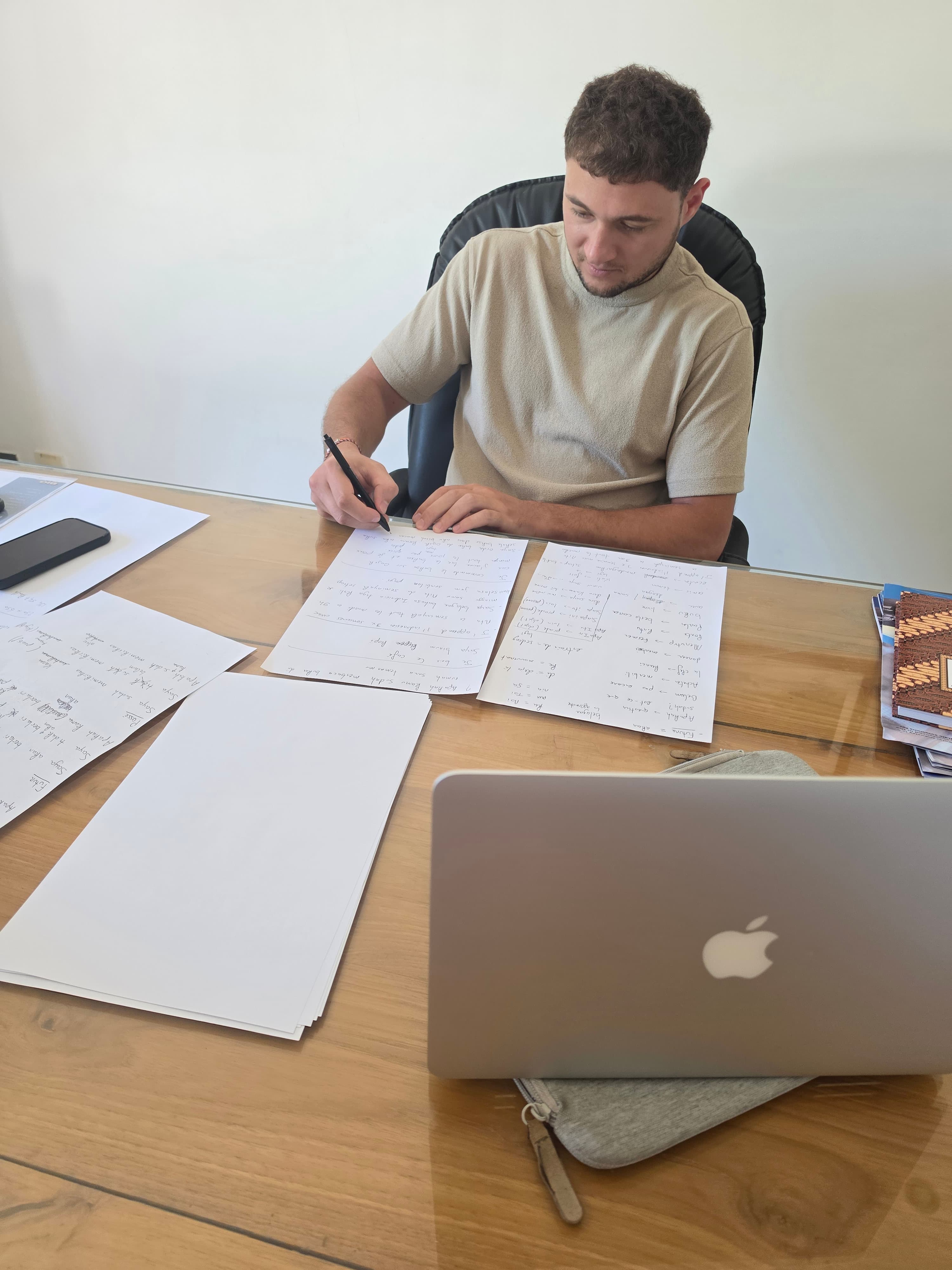 Jeune homme écrivant sur du papier à un bureau en bois avec un ordinateur portable.