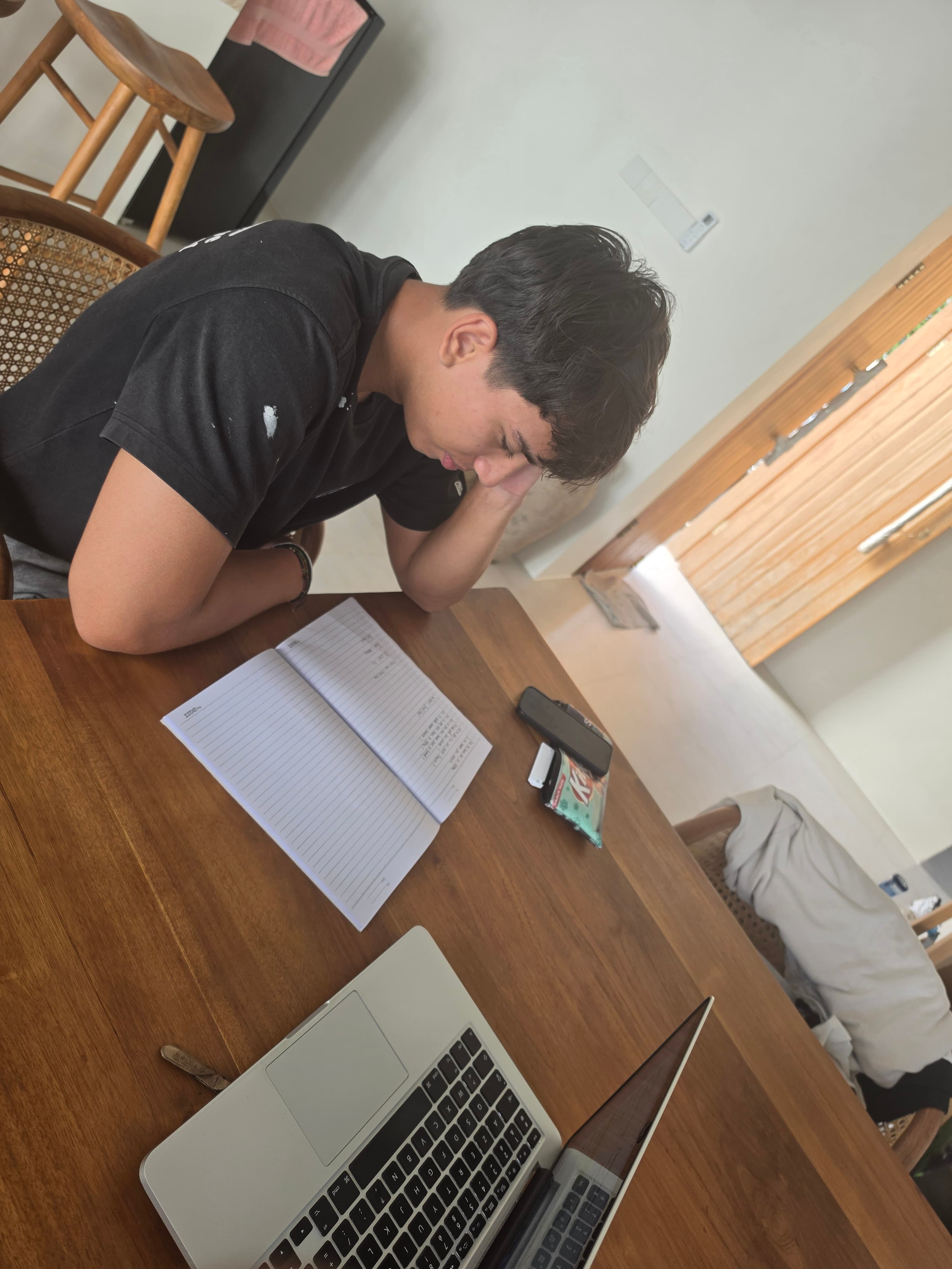 Jeune homme concentré sur son cahier avec un ordinateur portable sur une table en bois.