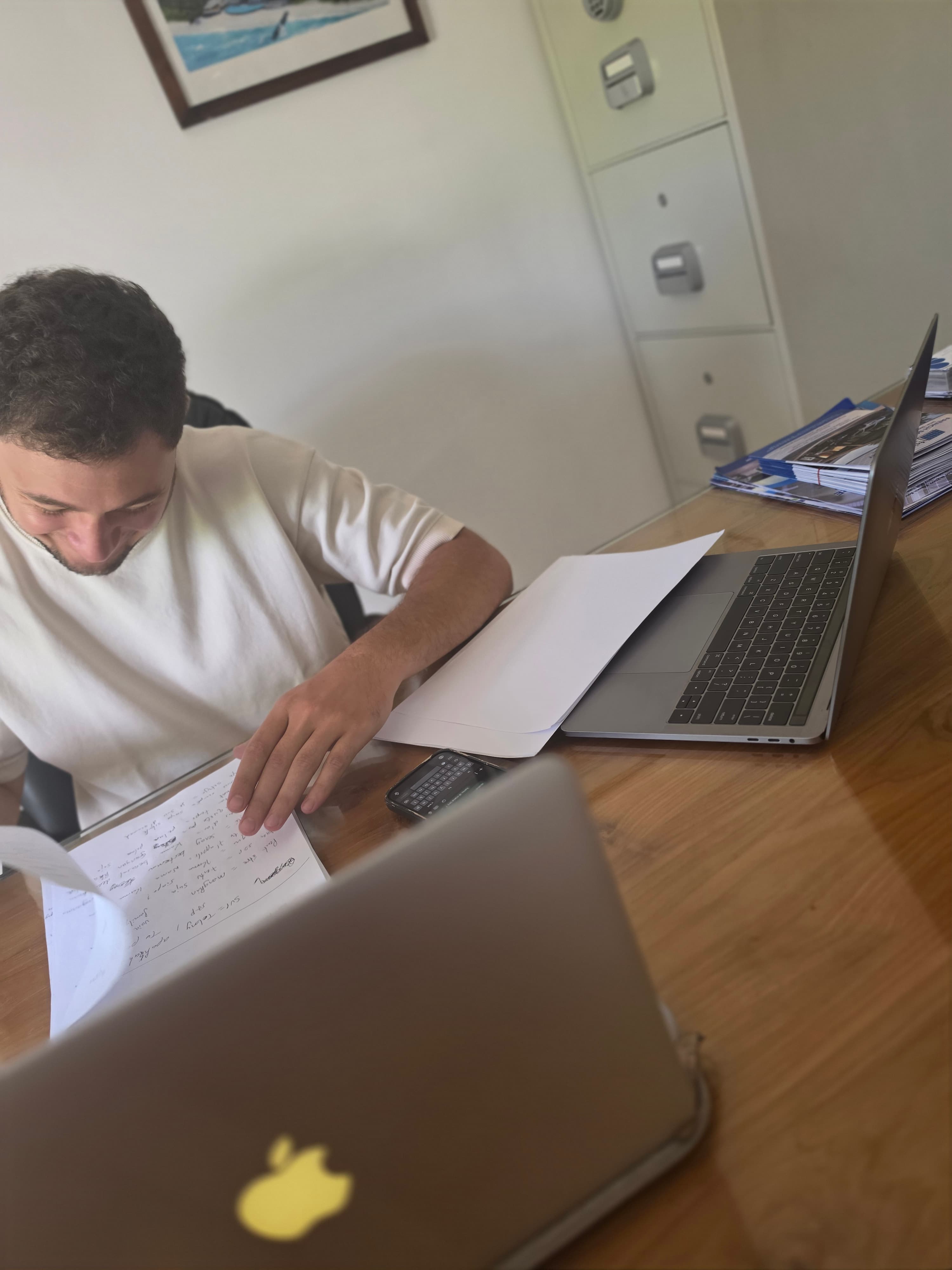 Homme en t-shirt blanc lisant des documents à son bureau avec un ordinateur portable.