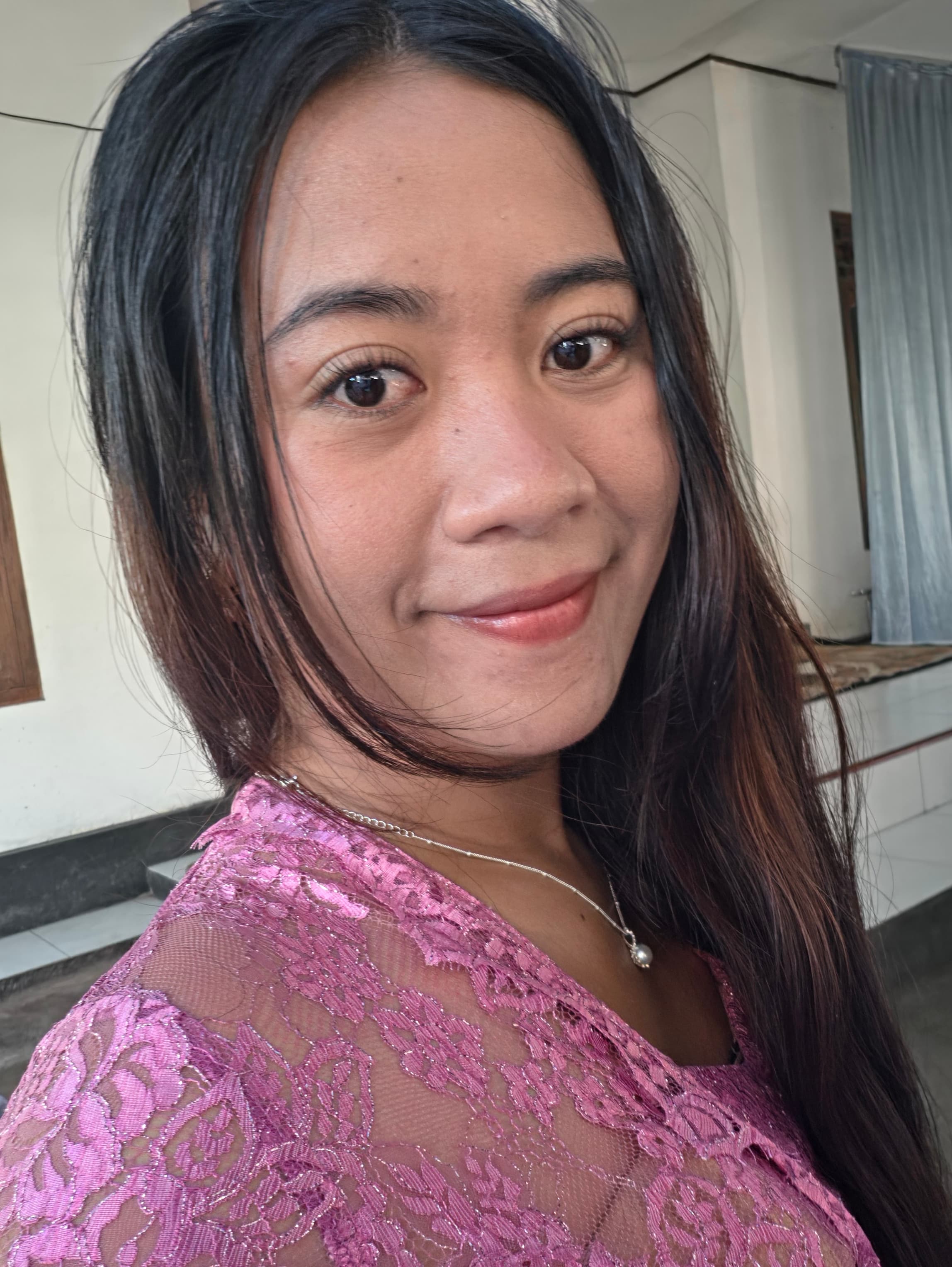 Femme souriante aux longs cheveux noirs, vêtue d'un haut en dentelle rose et d'un collier.